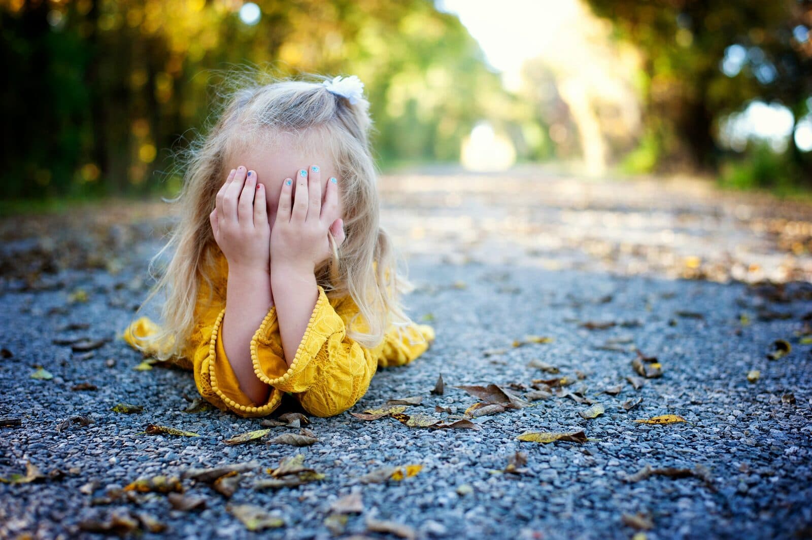 Young child in a yellow dress lying on an autumn path, hiding her face behind her hands